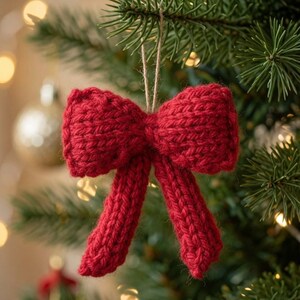 May include: A deep red knitted bow ornament featuring a textured yarn construction with two loops and two hanging tails. The bow is suspended by a thin, light-colored string loop, designed for hanging on a branch or similar support structure.
