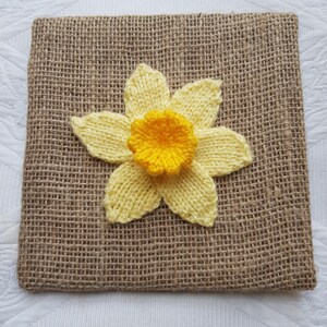 May include: A yellow knitted daffodil flower is attached to a brown hessian background.