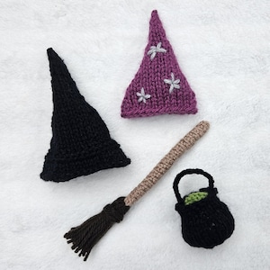 Könnte beinhalten: Ein Set aus gestrickten Miniaturartikeln, bestehend aus einem einfarbig schwarzen spitzen Hut, einem violetten spitzen Hut mit weißen gestickten Sternen, einem braunen Garnbesen und einem kleinen schwarzen Kessel mit grünem Innenfutter aus Garn.