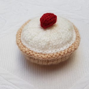 Könnte beinhalten: Ein gestrickter cremefarbener Kuchen mit einer roten gestrickten Kirsche oben drauf.