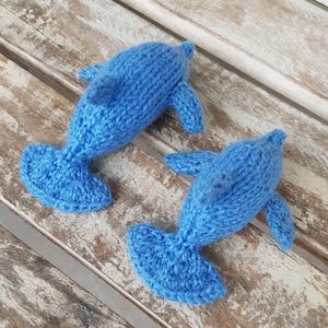 Könnte beinhalten: Zwei handgestrickte blaue Delfine. Jeder Delfin hat einen stromlinienförmigen Körper, Flossen und einen Schwanz. Die Delfine sind aus einem strukturierten Garn gefertigt, was ihnen ein weiches Aussehen verleiht. Der Hintergrund ist eine verwitterte Holzoberfläche.