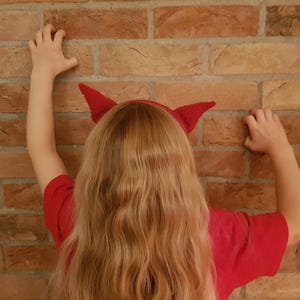May include: A red devil horn headband is worn by a person with long blonde hair, against a brick wall. The person is wearing a red shirt and has their arms raised, hands touching the wall.