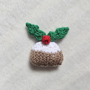 May include: A small knitted Christmas pudding ornament with a white and brown base and green holly leaves on top.
