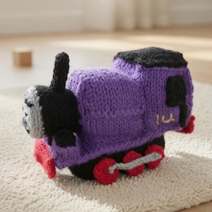 Könnte beinhalten: Ein handgestrickter Spielzeugeisenbahn aus violetter, schwarzer und roter Wolle. Der Zug hat ein hellgraues Gesicht mit gestickten schwarzen Details, einen schwarzen Schornstein, einen violetten Körper und rote Räder, die mit grauen Garnstangen verbunden sind. Die Zahl 1 ist in Beige gestickt.