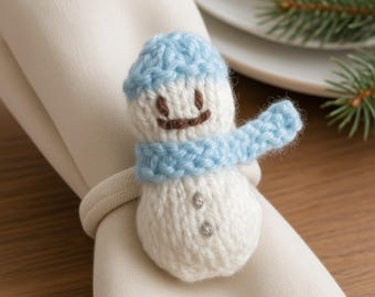May include: A white knitted snowman napkin ring featuring a light blue hat and scarf, brown embroidered eyes and mouth, and two small silver-colored buttons on the body, secured around a folded cream-colored fabric napkin.