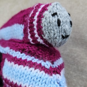 May include: A handmade knitted toy in the shape of a creature. The toy features a gray head with black stitched eyes and a smile. The body is a combination of burgundy, light blue, and white stripes. The knitted texture is clearly visible.