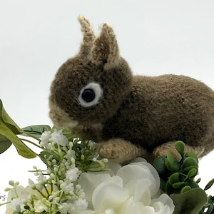 May include: A brown crocheted rabbit with white eyes and a white nose, sitting on a bed of white flowers and green leaves.
