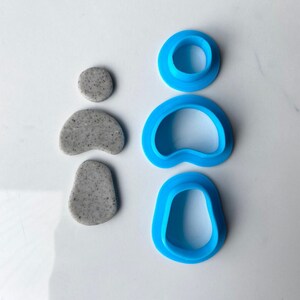 May include: Three blue plastic cookie cutters in the shape of a circle, a heart, and a teardrop. The cutters are on a white surface.