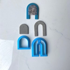 May include: A set of five blue plastic arch cookie cutters. The cutters are in different sizes and shapes. The largest cutter has three arches, the next largest has two arches, and the smallest cutter has one arch. There is also a small gray arch cookie cutter.