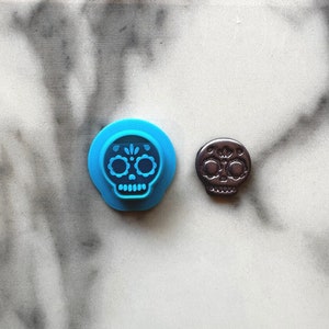 May include: Two sugar skull cookie cutters, one blue and one black. The blue cutter has a detailed design with white accents.