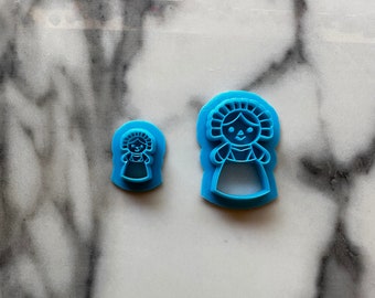 May include: Two blue plastic cookie cutters shaped like a traditional Mexican doll. The larger cutter is about 2 inches tall and the smaller cutter is about 1 inch tall.