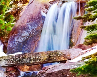 Könnte beinhalten: Ein Wasserfall stürzt über mit Moos bedeckte Felsen und eine Holzbrücke. Das Wasser ist milchig weiß und die Felsen sind tiefbraun und rot. Üppiges grünes Laub umgibt den Wasserfall.