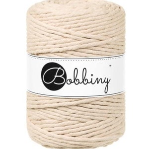 May include: A spool of natural-colored macrame cord. The cord is tightly wound and secured with a white label that reads "Bobbiny" in a cursive font. The cord appears to be made of cotton and is suitable for various crafting projects.