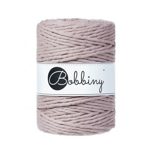 May include: A cylindrical spool of light mauve-colored macrame cord. A white label with the word "Bobbiny" in a script font is wrapped around the center of the spool. The cord is tightly wound, showing the texture of the material.