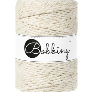 May include: A cylindrical spool of off-white macrame cord with gold metallic threads. The label on the spool reads "Bobbiny" in a cursive font within a black circle.