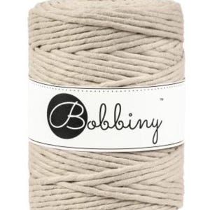May include: A spool of beige macrame cord with the brand name "Bobbiny" printed on a white label. The cord is tightly wound, showing a textured surface. Ideal for crafting projects.