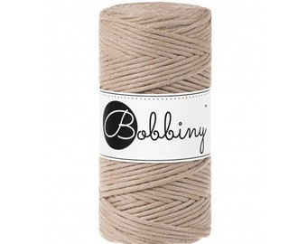 May include: A cylindrical spool of beige macrame cord. A white label with the word "Bobbing" in a script font is wrapped around the center of the spool. The cord is tightly wound, showing a textured surface.