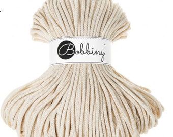 May include: A bundle of off-white macrame cord. The cord is tightly wound and secured with a white label that reads "Bobbiny" in a cursive font. The cord appears to be made of a soft, natural fiber, suitable for various crafting projects.
