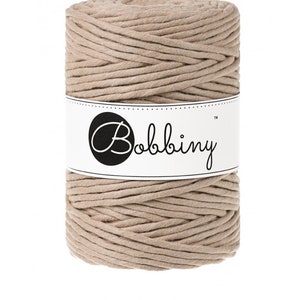 May include: A cylindrical spool of sand-colored Bobbiny macrame cord. The cord is tightly wound and wrapped around itself. A white label with the Bobbiny logo and the word "SAND" is visible.