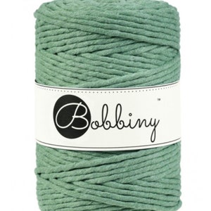 May include: A spool of sage green macrame cord. The cord is tightly wound and wrapped with a white label that reads "Bobbiny" in a black script font. The cord is a solid color and appears to be made of cotton.