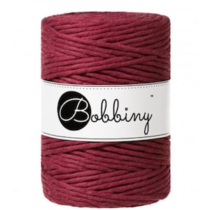 May include: A cylindrical spool of burgundy-colored macrame cord. A white label wraps around the center, featuring a black circle with the word "Bobbiny" in cursive script. The cord is tightly wound, showcasing a textured surface.