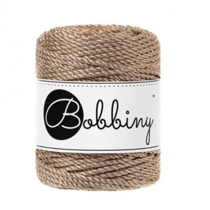 May include: A spool of natural-colored twine with the word "Bobbiny" printed on a white label. The twine is tightly wound, showing a textured surface. The label features a black circle with the brand name in a cursive font.