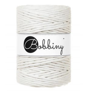 May include: A cylindrical spool of off-white braided cotton cord wrapped in a white paper band. The band features the brand name Bobbiny printed in a black script font inside a black circle on the left side, with a small trademark symbol.