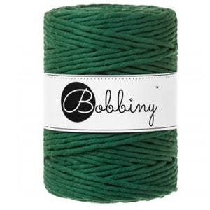 May include: A spool of dark green macrame cord. The cord is wrapped around itself in a cylindrical shape. A white label with the word "Bobbiny" in a script font is wrapped around the center of the spool.
