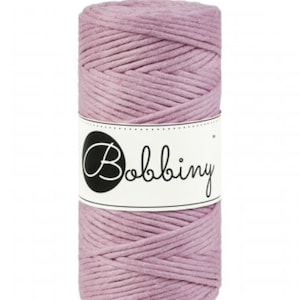 May include: A roll of mauve-colored macrame cord. The cord is tightly wound and wrapped with a white label featuring the word "Bobbiny" in a script font. The cord is suitable for various crafting projects.