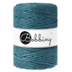 May include: A cylindrical spool of teal-colored macrame cord wrapped in a thick, braided texture. A white paper label is wrapped around the center of the spool, featuring the brand name Bobbiny printed in a black, stylized cursive font.