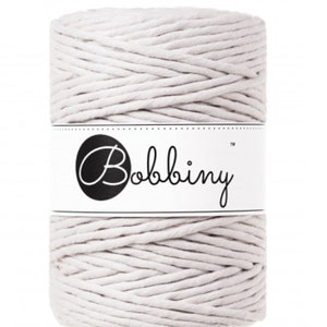 May include: A cylindrical spool of off-white macrame cord. The cord is tightly wound and wrapped with a white label featuring the brand name "Bobbiny" in a black circle. The cord appears to be made of a soft, natural fiber, suitable for various crafting projects.