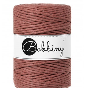 May include: A spool of dusty rose-colored macrame cord. The cord is wrapped around itself in a cylindrical shape. A white label with the word "Bobbiny" in a script font is wrapped around the center of the spool.