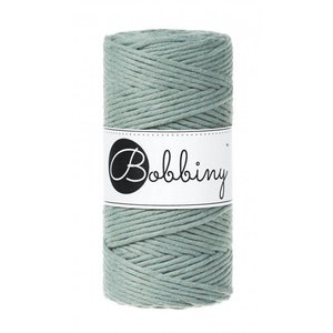 May include: A spool of sage green macrame cord. The cord is tightly wound and wrapped with a white label that reads "Bobbiny" in a black, elegant script. The cord appears to be made of a soft, woven material.