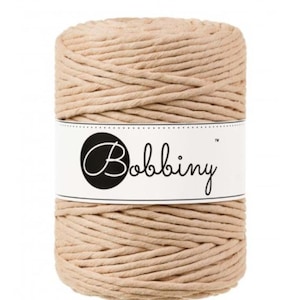 May include: A spool of beige macrame cord with the brand name "Bobbiny" printed on a white label. The cord is tightly wound, showing a textured surface. The label is wrapped around the center of the spool.