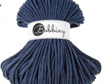 May include: A bundle of navy blue macrame cord. The cord is wrapped with a white label that reads "Bobbiny" in a cursive font. The cord is tightly wound and has a textured appearance.