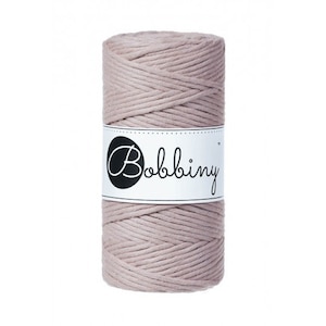 May include: A cylindrical spool of light taupe-colored macrame cord. The cord is wrapped around itself, creating a textured surface. A white label with the word "Bobbiny" in a script font is wrapped around the center of the spool.