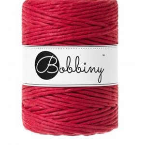 May include: A cylindrical spool of red yarn. The yarn is tightly wound and has a textured appearance. A white label with the word "Bobbiny" in a script font is wrapped around the center of the spool. The label has a black circle with a stylized "B" inside.
