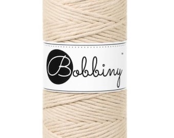 May include: A spool of natural-colored macrame cord. The cord is wrapped around itself, forming a cylindrical shape. A white label with the word "Bobbiny" in a script font is wrapped around the center of the spool. The background is plain white.