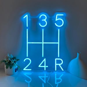 May include: A bright blue neon sign displaying a manual transmission gear shift pattern. The layout features the numbers 1, 3, 5 across the top and 2, 4, R across the bottom, connected by vertical and horizontal lines forming the shift gate.