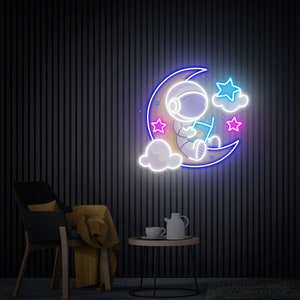 May include: Neon sign depicting an astronaut sitting on a crescent moon with clouds and stars. The astronaut is reading a book. The sign is lit in blue and white.