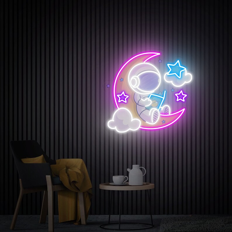 May include: Neon sign depicting an astronaut sitting on a crescent moon reading a book. The moon is pink and the astronaut is white. There are white clouds and blue stars around the moon.