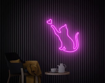 May include: Pink neon sign depicting a cat reaching up to catch a heart. The sign is mounted on a black wall with a chair and a small table in the foreground.