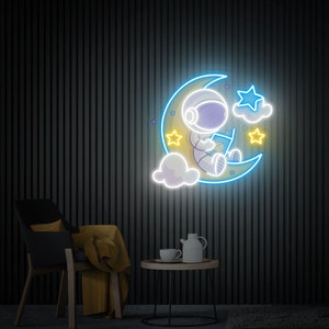 May include: A neon sign depicting an astronaut sitting on a crescent moon with stars and clouds. The astronaut is holding a book and the moon is yellow with a blue outline. The sign is on a black wall.