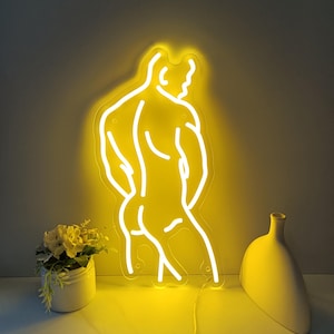 Puede incluir: Una luz de neón amarilla con la forma de una figura musculosa, vista desde atrás. La luz de neón está montada en una pared. Un pequeño jarrón blanco y una pequeña maceta con flores están en primer plano.