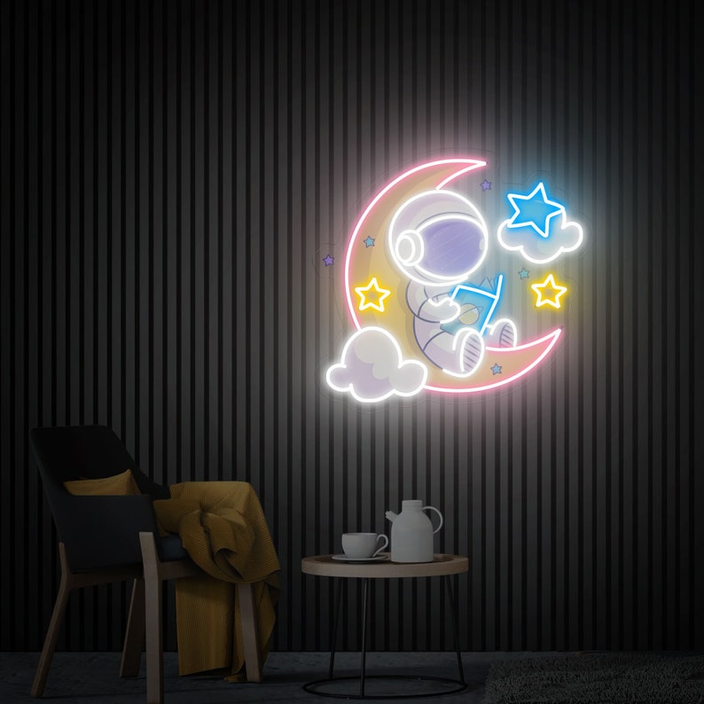 May include: Neon sign depicting an astronaut sitting on a crescent moon, reading a book. The moon is pink and yellow, with white clouds and yellow stars. The astronaut is wearing a white spacesuit with a blue helmet.