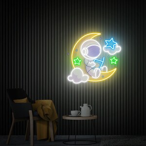 May include: Neon sign depicting an astronaut sitting on a crescent moon, reading a book. The moon is yellow, the astronaut is white with blue accents, and the stars are green and blue.