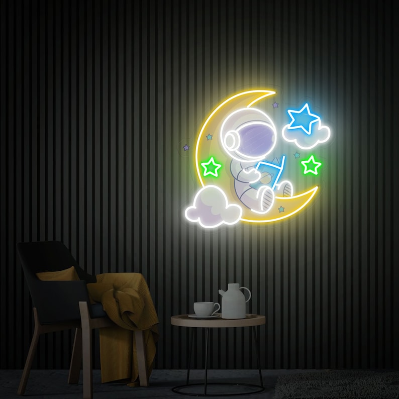 May include: Neon sign depicting an astronaut sitting on a crescent moon, reading a book. The moon is yellow, the astronaut is white with blue accents, and the stars are green and blue.