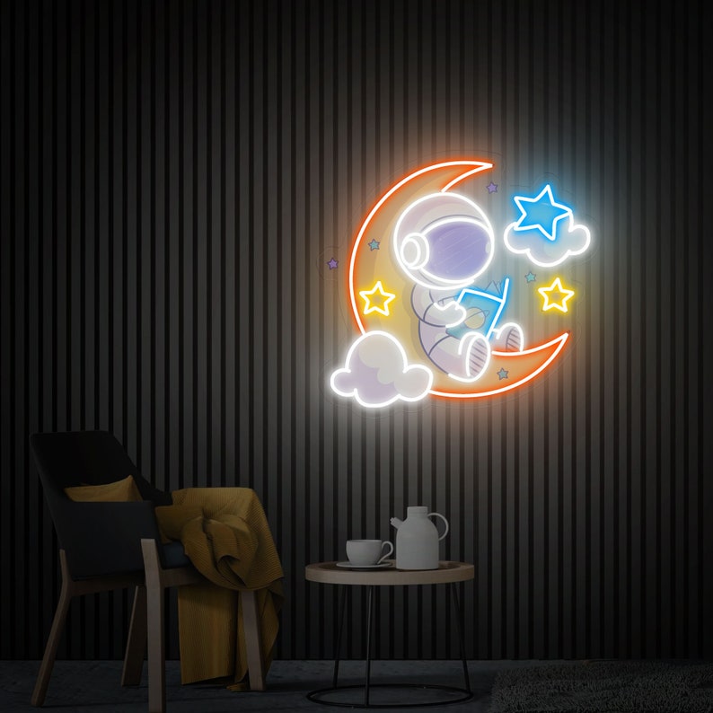 May include: Neon sign depicting an astronaut sitting on a crescent moon, reading a book. The moon is orange and yellow, the astronaut is blue and white, and the stars are yellow and blue.