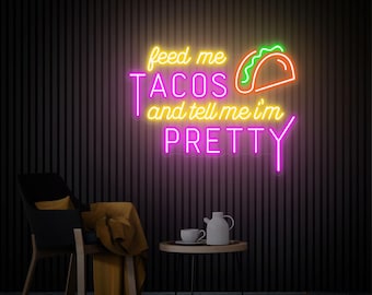 May include: A neon sign with the text "feed me TACOS and tell me I'm PRETTY" in yellow, pink, and orange. The sign features a taco graphic. The sign is mounted on a dark, vertically-lined wall. A chair, table, and rug are in the foreground.