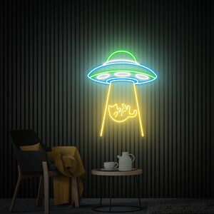 May include: A neon sign depicting a UFO in green, blue, and white, beaming a yellow cat. The sign is mounted on a dark, vertically striped wall. A black chair with a yellow blanket and a small table with a teapot and cup are in the foreground.
